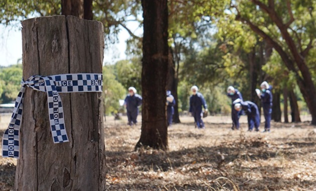 With police tape in foreground, a group of students in boiler suits walk though forest and investigate a mock-crime scene