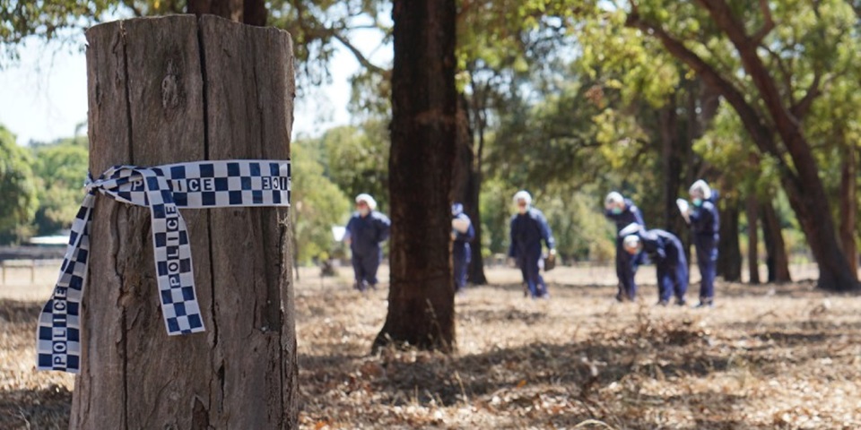 With police tape in foreground, a group of students in boiler suits walk though forest and investigate a mock-crime scene