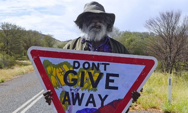 Iltja Ntjarra artist Mervyn Rubuntja holding the Don't Give Away hand-painted sign.