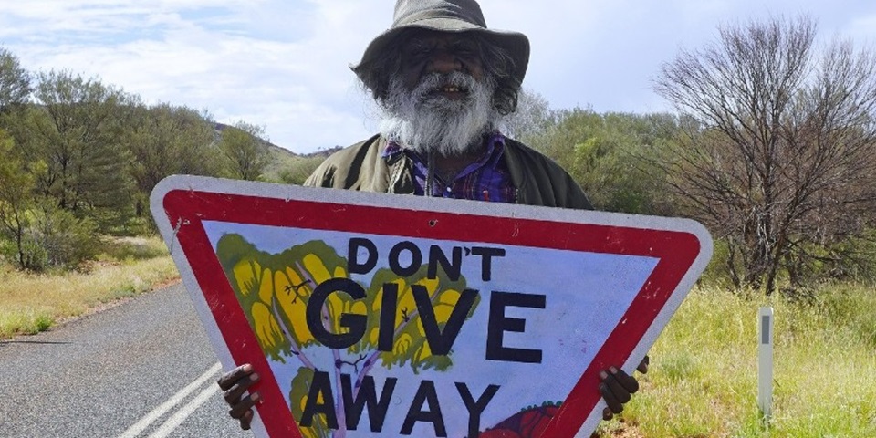 Dont_Give_Away_feature Iltja Ntjarra artist Mervyn Rubuntja holding the Don't Give Away hand-painted sign.