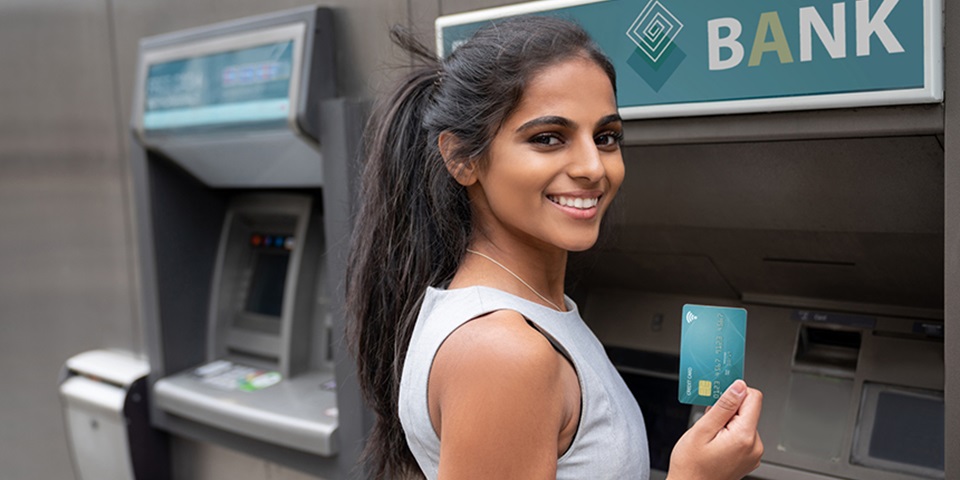 How to open a bank account feature Female student smiling in front of ATM holding up a debit card