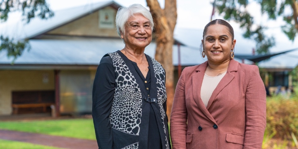 Two women smiling, looking at the camera, in front of a building shrouded in lush green native trees