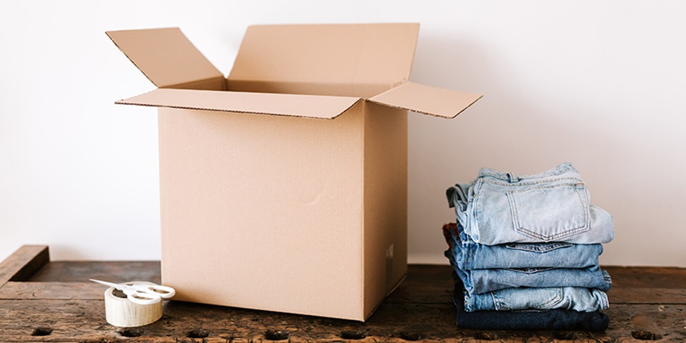 Moving to the city feature Folded jeans next to an open cardboard box on a wooden table