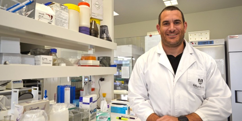 man with brown hair in a lab wearing a lab coat and smiling at the camera