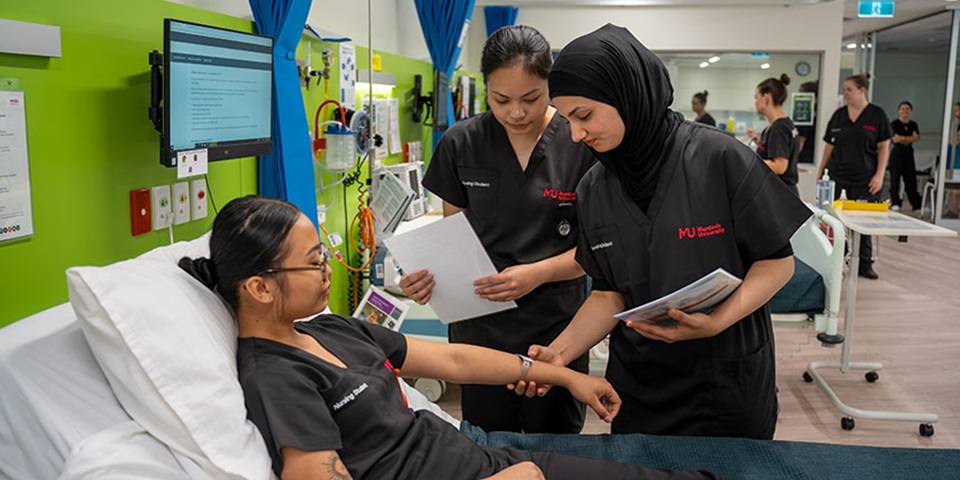 Nursing_Job_Feature Two female students take the pulse of a third student who is lying on a bed in the SimLab.