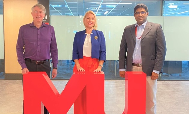 Dr James Trotter, Monika Fourneaux and Joseph John Nalloor standing behind a large MU sign