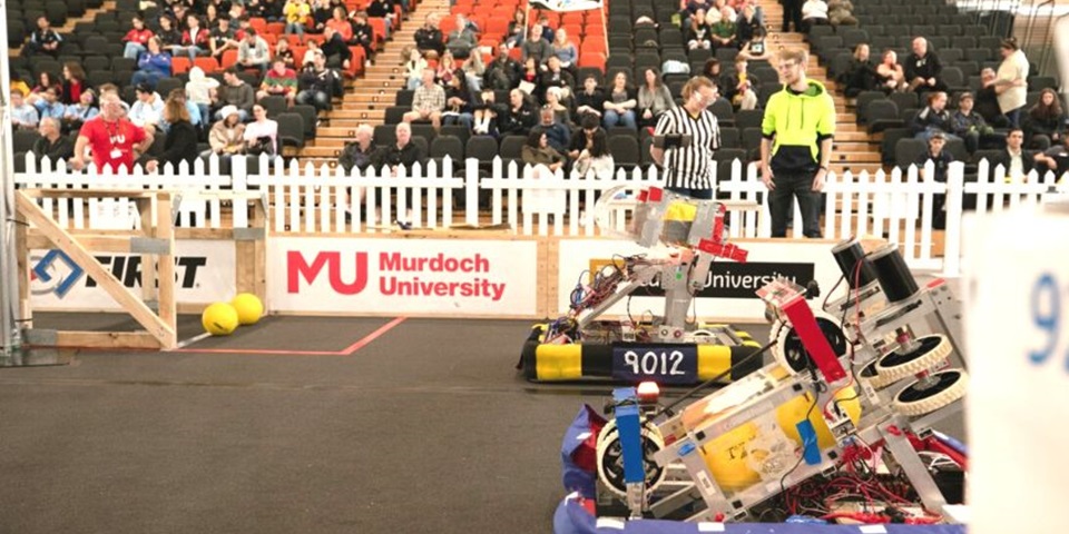 Two robots in the foreground in an indoor fenced area with stadium seats behind the short fence. Two people inside the fence with the robots and spectators in the stands.