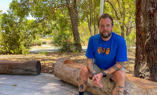 Man sitting on tree log in bush setting looking towards camera