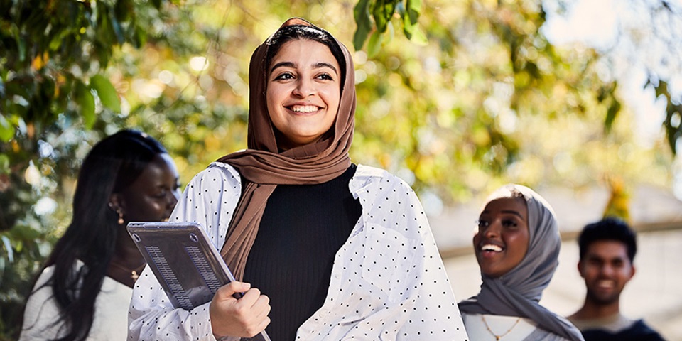 Social_Study_Feature Female student wearing hijab smiling and holding laptop.