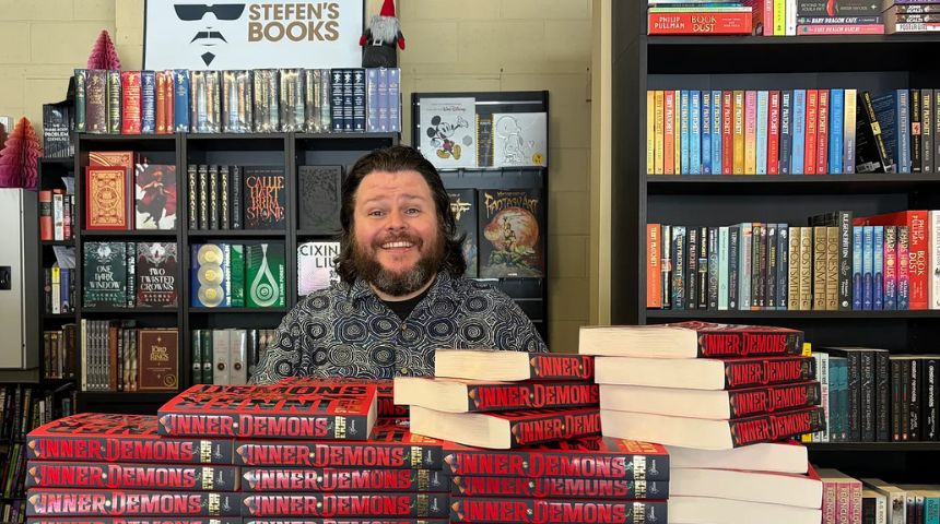 Stephen is standing in Stefen's Bookshop in front of a pile of his novels, smiling.